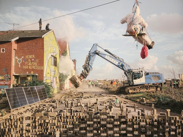 2023년 11월 뤼체라트 마을의 강제 퇴거 첫 날 한 기후환경운동가가 구조물에 매달려 진압 속도를 늦추는 동안 굴착기가 운동가들이 설치한 바리케이트를 제거하고 있다. 